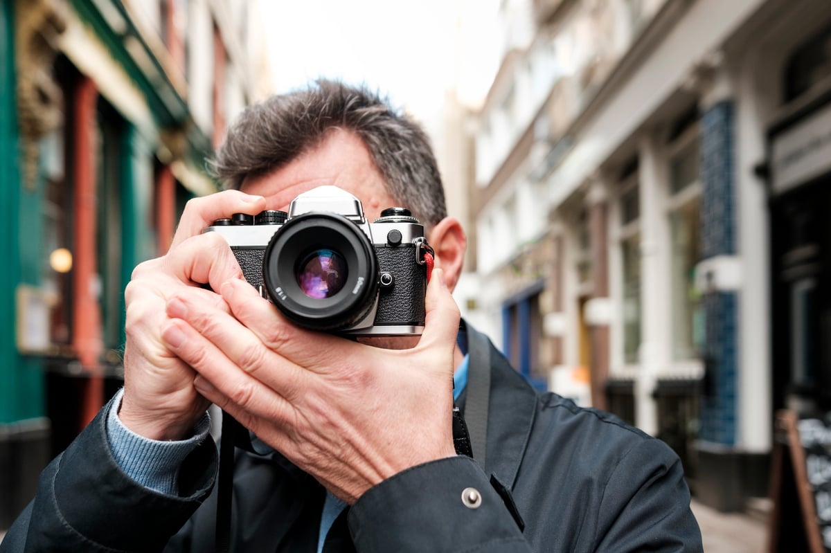 Mature male photographer with gray hair using a vintage film camera on a european city street, capturing urban architecture and candid travel moments during a daytime stroll