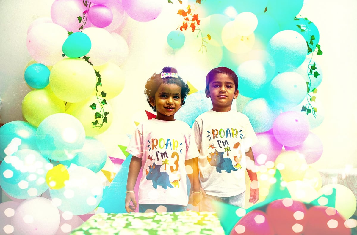 Two young children wearing matching graphic t-shirts stand in front of a colorful balloon backdrop with confetti