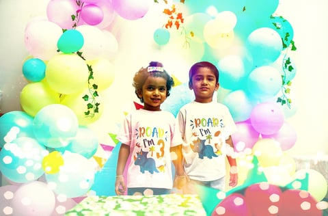 Two young children wearing matching graphic t-shirts stand in front of a colorful balloon backdrop with confetti