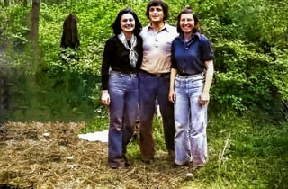 Three women standing together outdoors in a forest setting with green foliage in the background